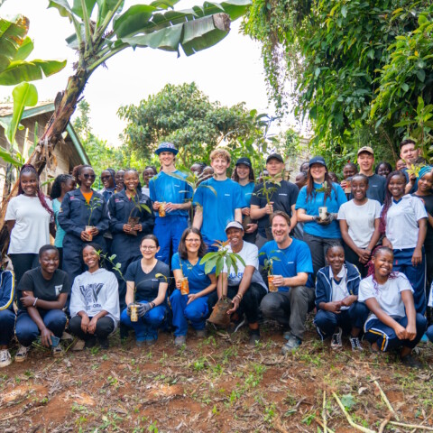 A group of people stands together in a lush outdoor setting, smiling and holding small plants, with trees and greenery in the background.