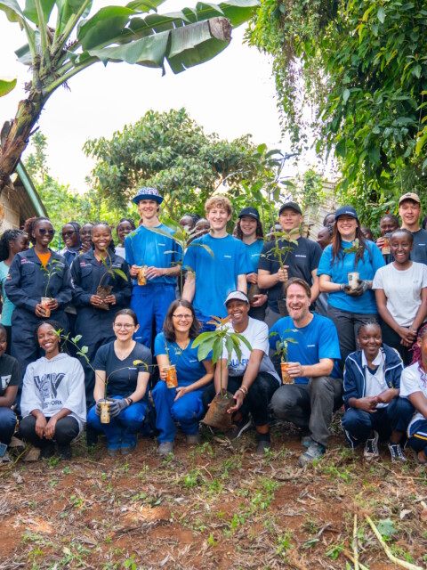 A group of people stands together in a lush outdoor setting, smiling and holding small plants, with trees and greenery in the background.
