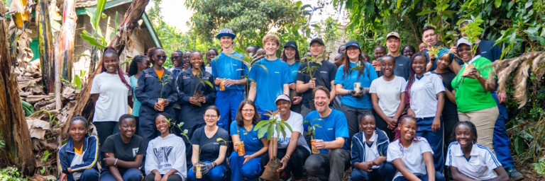 A group of people stands together in a lush outdoor setting, smiling and holding small plants, with trees and greenery in the background.