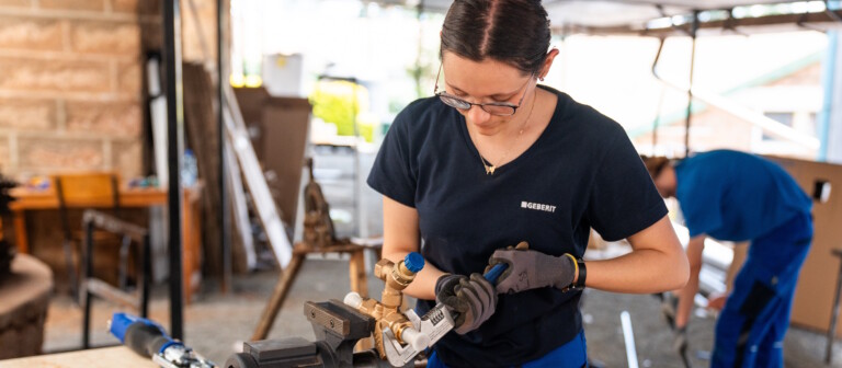 A woman in a workshop is working with plumbing components, using tools and a vice on a table.