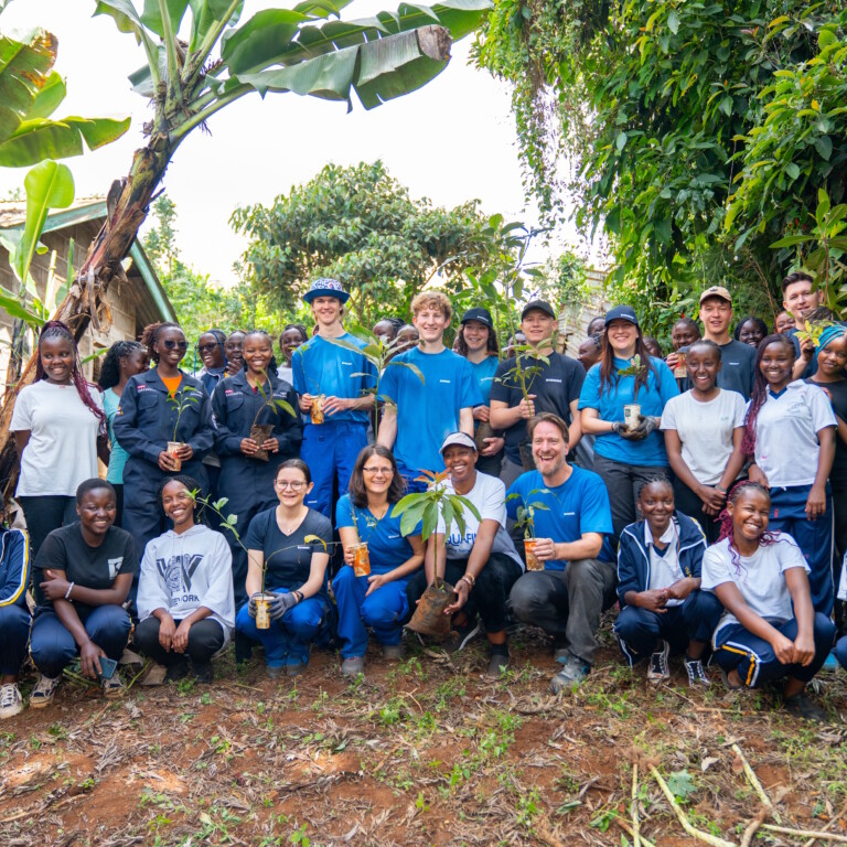 A group of people stands together in a lush outdoor setting, smiling and holding small plants, with trees and greenery in the background.