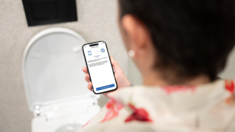 A person holding a smartphone displaying an app interface while facing a modern toilet.