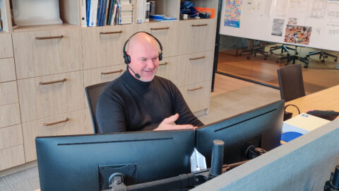 A man wearing a headset is sitting at a desk with two computer monitors, smiling and gesturing.