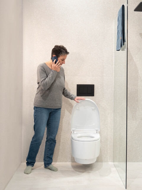 A person standing next to a modern toilet in a bathroom, talking on the phone.