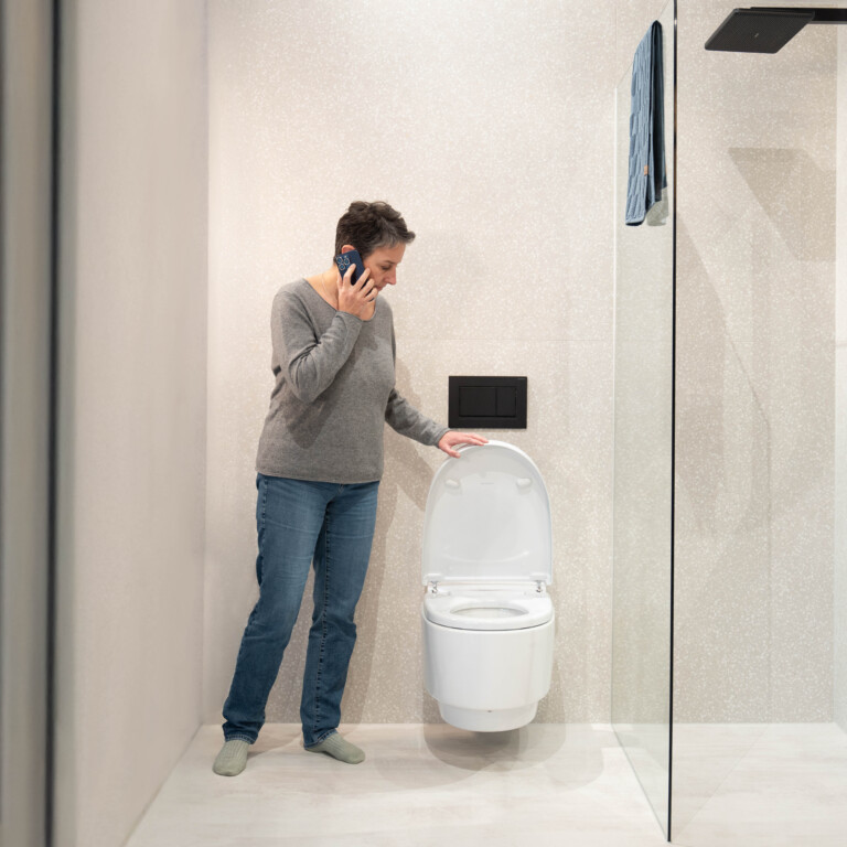 A person standing next to a modern toilet in a bathroom, talking on the phone.