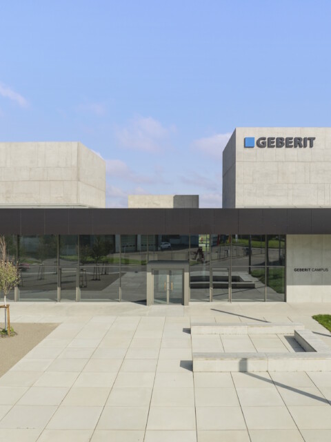 Moderne Unternehmensarchitektur mit betonsichtbaren Würfeln und Glasfassade, begrüntem Areal und gepflastertem Eingang.