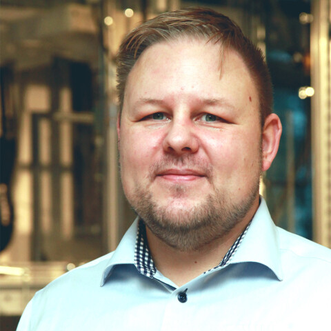 Ein Mann mit kurzen, hellen Haaren und einem Bart, der ein hellblaues Hemd trägt, steht vor einer unscharfen Hintergrundszene.