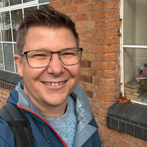 Lächelnde Person mit Brille, trägt eine Jacke und steht vor einer Backsteinwand mit Fenstern.