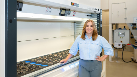 Eine Frau mit hellblauer Bluse steht neben einer Maschine in einer industriellen Umgebung.