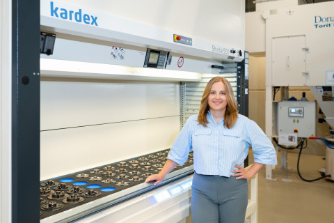 A woman stands next to a large industrial storage system, smiling and wearing a light blue blouse.