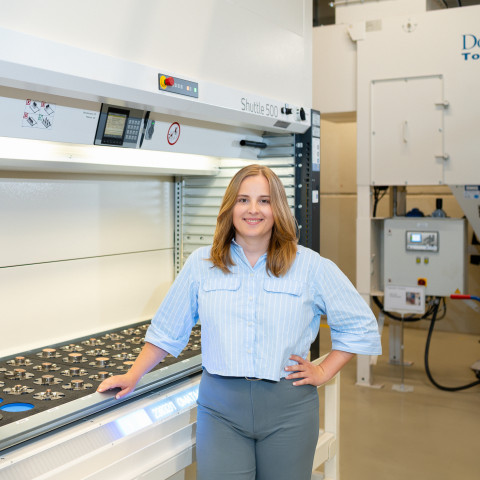 Eine Frau mit hellblauer Bluse steht neben einer Maschine in einer industriellen Umgebung.