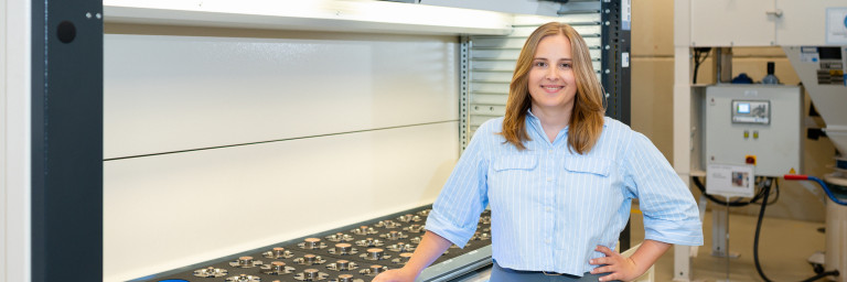 Eine Frau mit hellblauer Bluse steht neben einer Maschine in einer industriellen Umgebung.