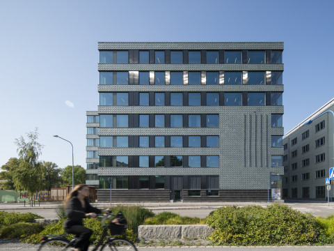 Modern building with geometric facade design, surrounded by greenery and a cyclist passing by.