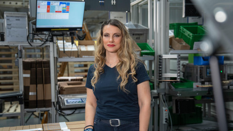 Eine Person mit langen, lockigen Haaren steht in einer Industrieumgebung mit Kisten und einem Bildschirm im Hintergrund.