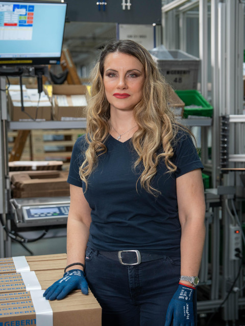 A woman with long, wavy hair stands in a warehouse environment, wearing a blue shirt and gloves, with boxes and a computer display in the background.