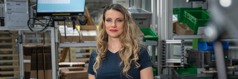 A woman with long, wavy hair stands in a warehouse environment, wearing a blue shirt and gloves, with boxes and a computer display in the background.