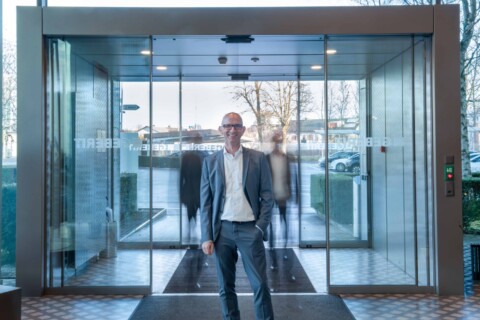 A man in a gray suit stands at the entrance of a modern glass building with automatic sliding doors, with blurry silhouettes in the background.