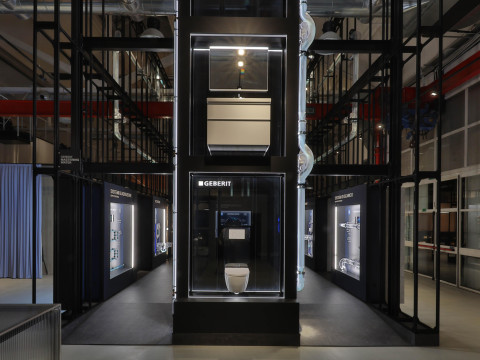 A modern, industrial space featuring a tall, black multi-level structure with glass panels and illuminated sections, surrounded by an open area.