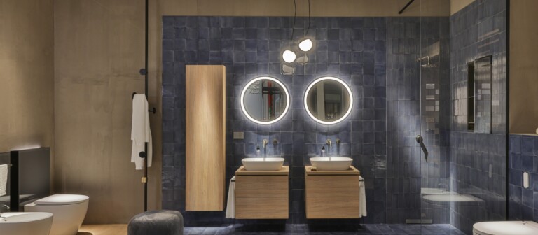 Modern bathroom featuring dual wooden sink cabinets, round backlit mirrors, a shower area, and stylish fixtures against a blue tile backdrop.