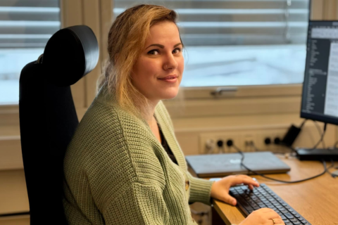 A young woman with blonde hair sits at a desk in an office, smiling while using a computer.