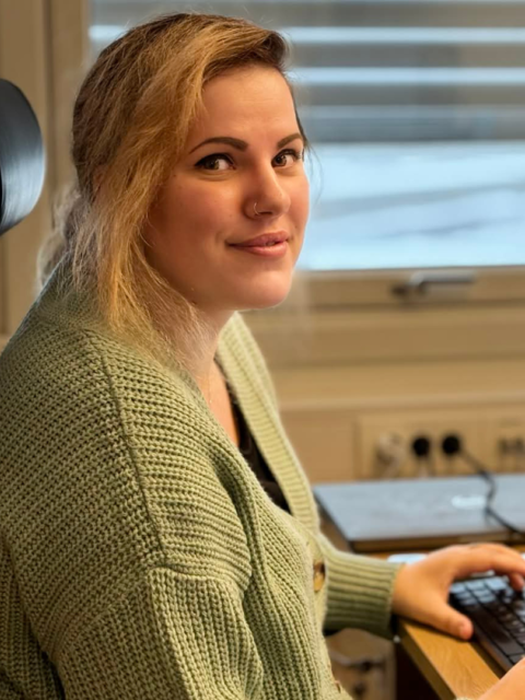 A young woman with blonde hair sits at a desk in an office, smiling while using a computer.