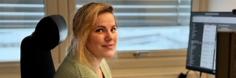 A young woman with blonde hair sits at a desk in an office, smiling while using a computer.