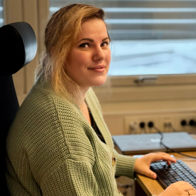 A young woman with blonde hair sits at a desk in an office, smiling while using a computer.