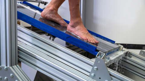 A person's bare foot on a blue ramp, part of a testing apparatus with visible metal support structures.