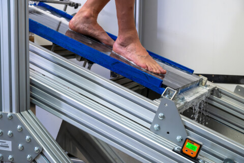 A person's bare foot on a blue ramp, part of a testing apparatus with visible metal support structures.