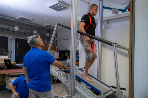 A person wearing a safety harness walks along a sloping surface with water, whilst others assist and provide support.
