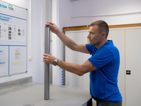 A man in a blue shirt is holding a metal cylinder and a steel ball above a shower tray.