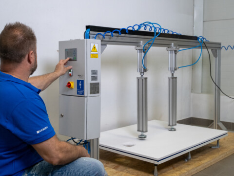 A man in a blue shirt operates a control panel for a testing machine with vertical adjustable columns in a workshop setting.