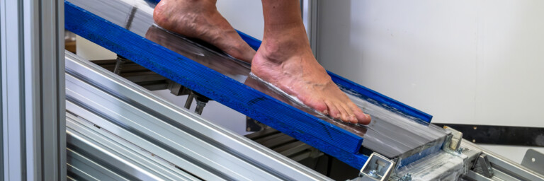A person's bare foot on a blue ramp, part of a testing apparatus with visible metal support structures.