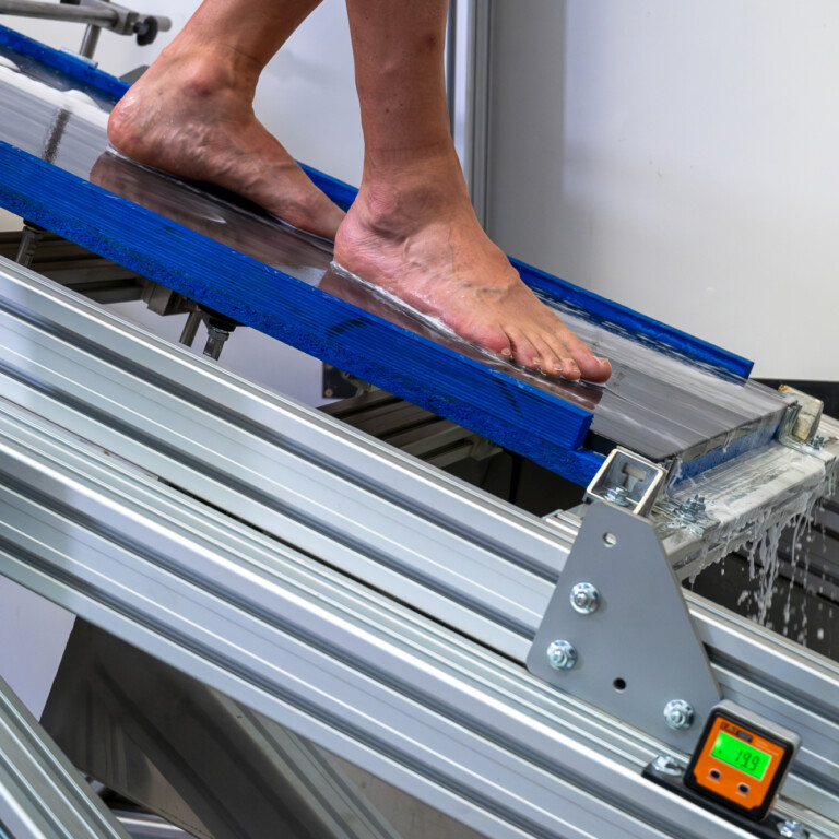 A person's bare foot on a blue ramp, part of a testing apparatus with visible metal support structures.