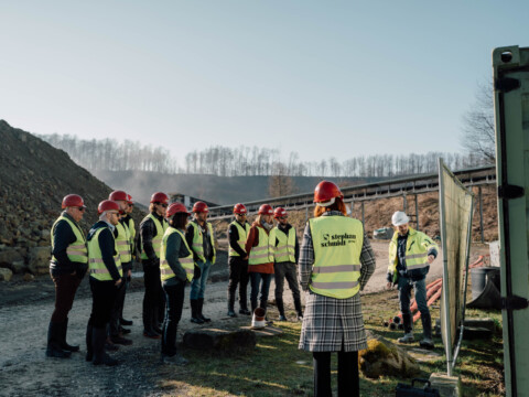 Eine Gruppe von Leuten, die Schutzkleidung tragen, darunter Helme und Warnwesten, hat sich versammelt, und einer von ihnen gibt Erläuterungen.