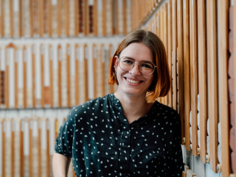 Eine lächelnde Frau mit kurzen, Haaren und Brille steht neben einer holzverkleideten Wand mit einem Muster aus vertikalen Streifen.