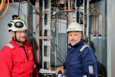 Zwei Männer in Arbeitskleidung und Schutzhelmen stehen vor einer technischen Anlage mit Rohren und Kabeln.