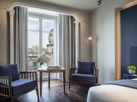 Ein elegantes Hotelzimmer mit zwei blauen Stühlen, einem Tisch und grossen Fenstern, die auf eine Stadtansicht blicken.