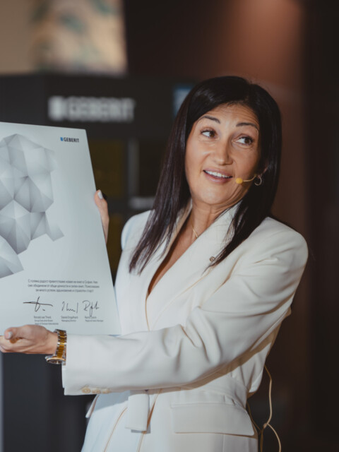 A woman in a white blazer holds up a map with a geometric design, smiling and presenting in a decorated space with balloons.