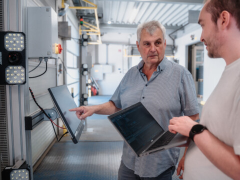 Two men engaged in a discussion, one holding a laptop and pointing toward equipment.