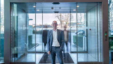 Ein Mann in einem grauen Anzug steht vor einer automatischen Glastür, während unscharfe Silhouetten von Personen vorbeigehen.