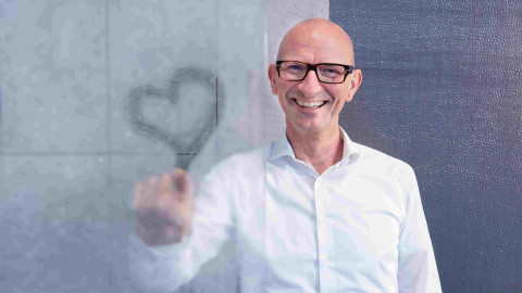 A smiling man with glasses is drawing a heart on a foggy glass wall.