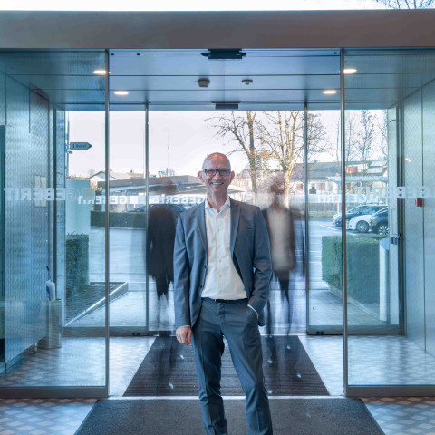 A man in a gray suit stands at the entrance of a modern glass building with automatic sliding doors, with blurry silhouettes in the background.
