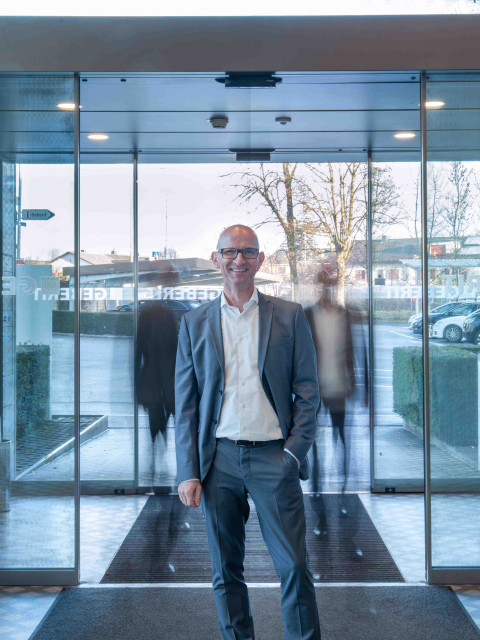 Ein Mann in einem grauen Anzug steht vor einer automatischen Glastür, während unscharfe Silhouetten von Personen vorbeigehen.