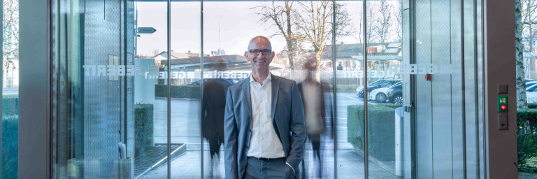 A man in a gray suit stands at the entrance of a modern glass building with automatic sliding doors, with blurry silhouettes in the background.