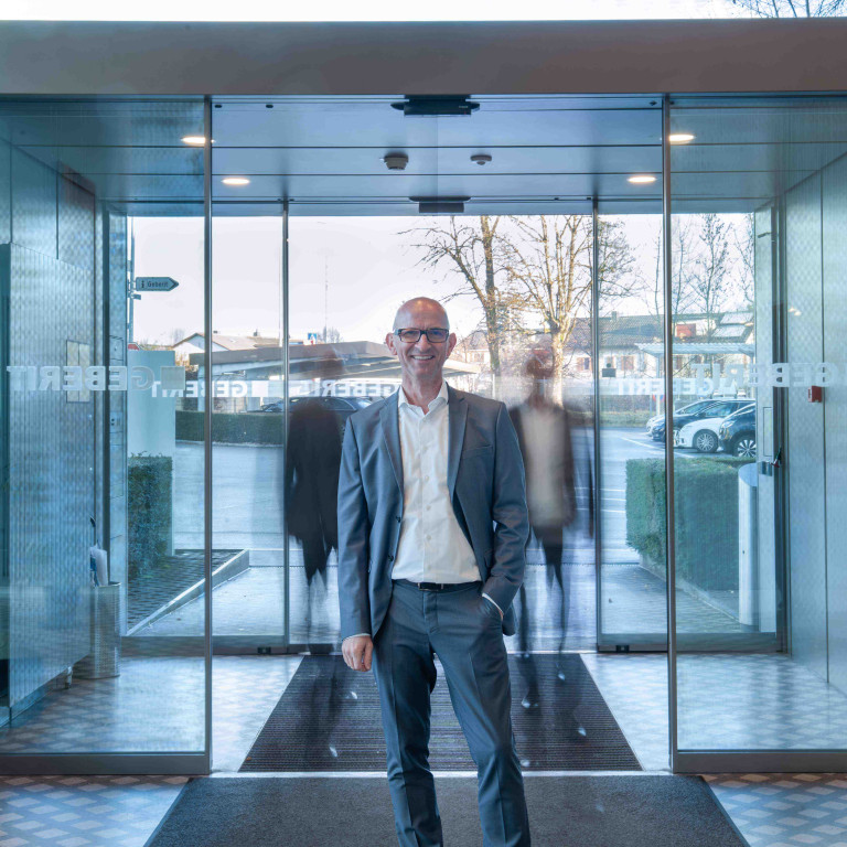 A man in a gray suit stands at the entrance of a modern glass building with automatic sliding doors, with blurry silhouettes in the background.