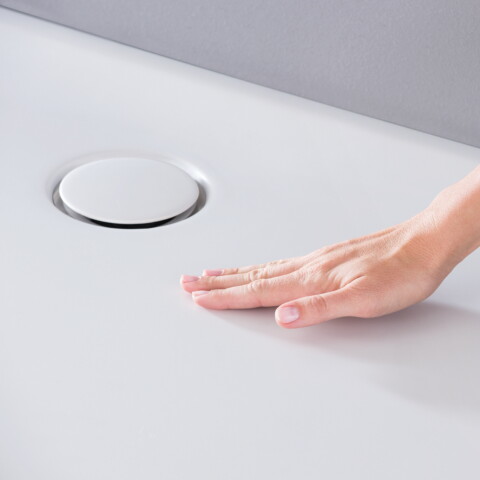 Eine Hand, die eine Duschwanne aus weissem Acryl mit einem kreisförmigen Abfluss berührt.