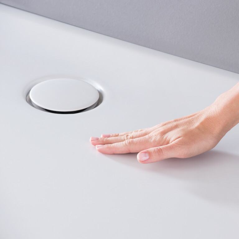 Eine Hand, die eine Duschwanne aus weissem Acryl mit einem kreisförmigen Abfluss berührt.