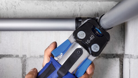 A hand operating a pressing tool on a pipe, connecting two sections of plumbing against a brick wall.