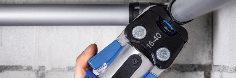 Eine Hand hält ein Presswerkzeug, das zwei an einer weiss gestrichenen Ziegelwand verlaufende Rohre verbindet.